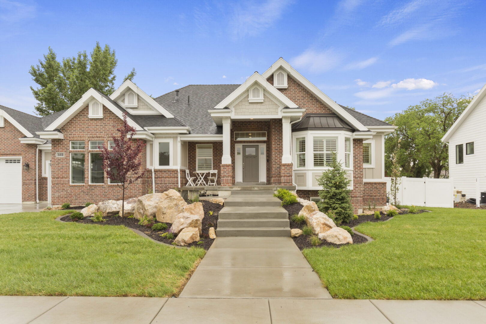A single-story brick house with a gabled roof, front porch, and landscaped yard featuring rocks and small trees. A concrete walkway leads to the entrance.